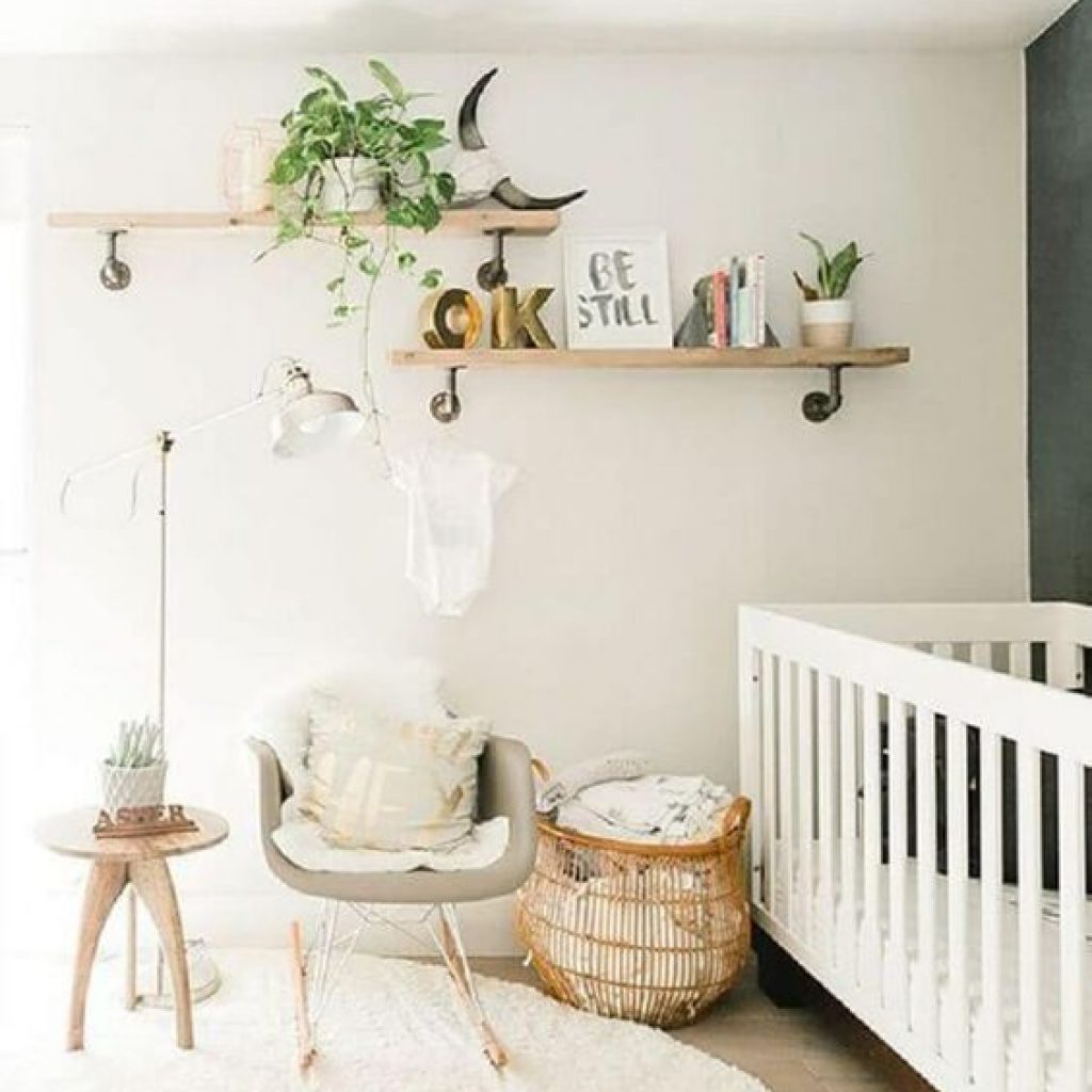Quarto de bebê com um berço branco, uma parede escura e outra branca, com uma prateleira e folhas naturais. Uma cadeira de balanço, um cesto e uma cabeceira.