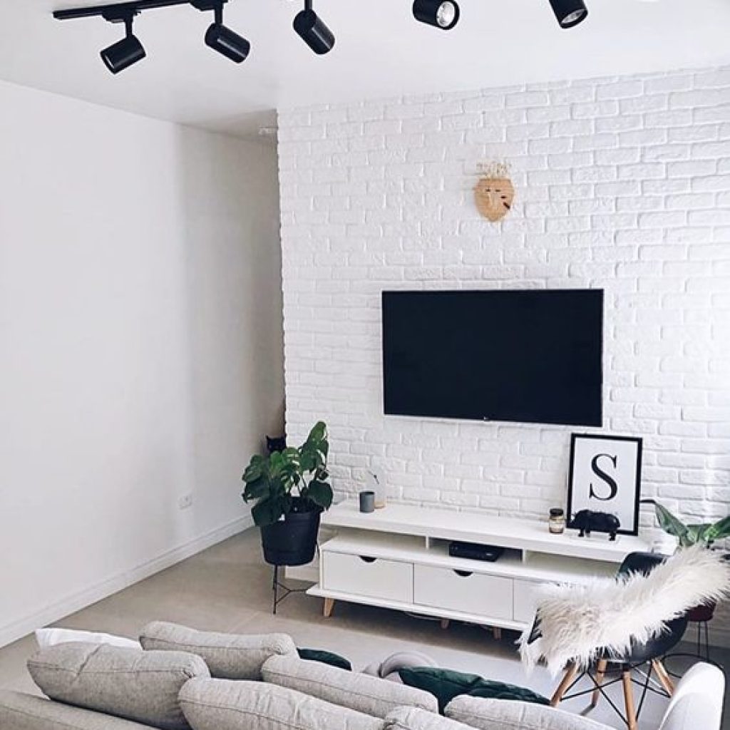 Sala com parede branca e de tijolinho. Há uma televisão pregada na parede e um sofá cinza.