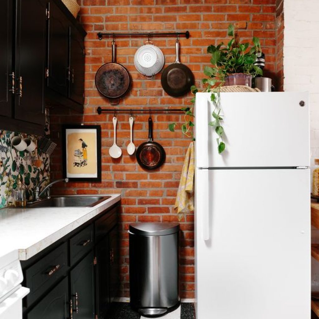 Cozinha com tijolinho à mostra na parede, armários pretos, panelas penduradas na parede, uma geladeira antiga branca, com um vaso de folhas em cima.
